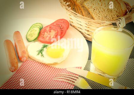 Le petit-déjeuner sur une table, œuf frit dans une saucisses frites en forme de cœur, des tranches de légumes concombres et tomates, jus, pain de mie, couteau et fourchette, Banque D'Images