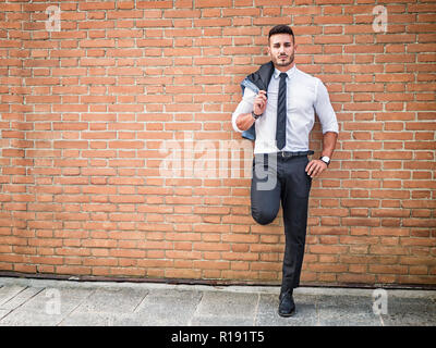 Portrait de jeune homme élégant costume d'affaires port, debout dans la ville moderne, appuyé contre un mur de briques Banque D'Images