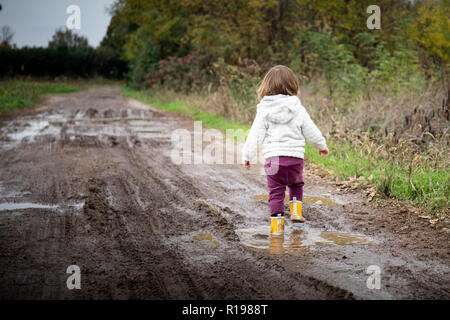 Bébé fille, vue de dos, les projections dans les flaques boueuses en route de campagne avec des bottes de pluie jaune. Copie de gauche l'espace. Banque D'Images