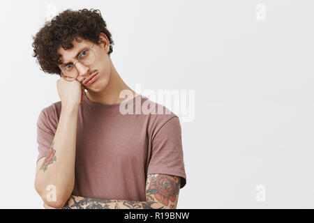 Belle et élégante jeune homme avec moustache cheveux frisés et bras tatoués leaning on fist tacaud et bouder les sourcils tandis qu'offensé d'être en colère contre un Banque D'Images