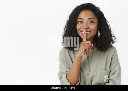 Chut, pouvez-vous garder de secrets. Portrait de charmant friendly-ravi à la femme à la peau sombre mignon avec curly hairstyle disant chut et souriant prepar Banque D'Images