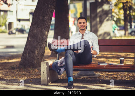 Un bel homme d'affaires est en plein air, à la recherche tandis que satisfait assis sur un banc, il est relaxant et profiter de Banque D'Images