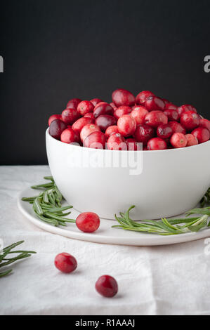 La canneberge rouge festif dans un bol blanc sur une plaque recouverte de feuilles de romarin. Banque D'Images