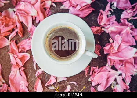 Mise à plat tasse de café avec motif en forme de coeur entouré de pétales de fleurs de pivoine rose clair sur fond noir en noir. Valentines Day, bonjour Banque D'Images