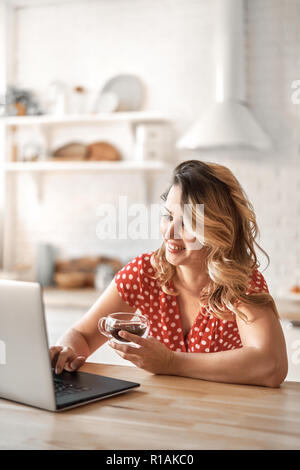 Heureux belle femme de boire du café. Femme au travail sur un ordinateur portable dans la cuisine. Banque D'Images