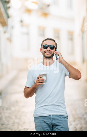 Bel homme vêtu de vêtements branchés en vous promenant sur la rue de la ville d'avoir une conversation téléphonique à l'aide d'opérateur international et d'itinérance holding coffee pour aller Banque D'Images