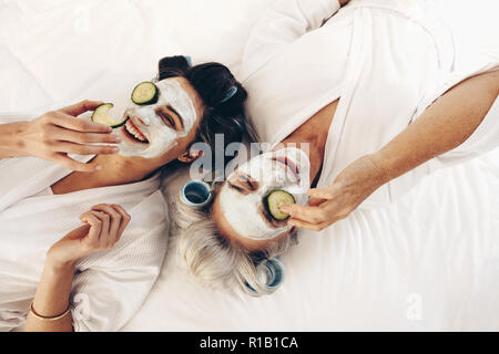 Deux femmes en peignoirs lying on bed wearing beauty face pack avec des tranches de concombres. Mère et fille se détendre sur le lit dont le visage pack et le rouleau cu Banque D'Images