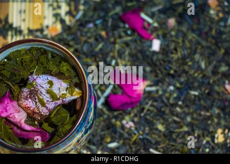 Thé vert parfumé aux pétales de rose rouge brassée dans une tasse avec des ornements de la culture chinoise. Une tasse se dresse contre l'arrière-plan de thé séchées éparses Banque D'Images