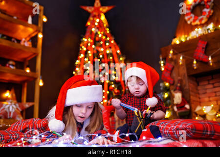 Fille charmante à Santa hat se trouvant sur le plancher et écrit au crayon, dessine la lettre et son petit frère qui interfère avec elle, joue avec des crayons dans une chambre chaude de guirlandes, de lumières Banque D'Images