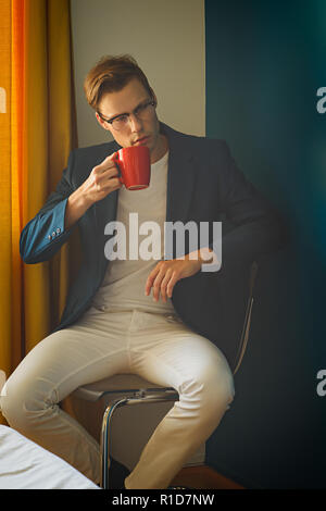 Un jeune homme, modèle masculin en costume bleu et blanc pantalon, porte une paire de lunettes vintage, à l'écart,tenant une tasse rouge. Un men's vintage fashion concept. Banque D'Images