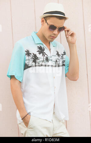 Un beau modèle masculin posant dans des vêtements d'été piscine contre un mur en bois. Il porte un chapeau fedora blanc et aviator sunglasses. Banque D'Images