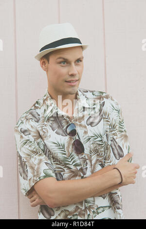 Un beau modèle masculin posant dans des vêtements d'été en plein air avant d'un mur en bois clair. Il porte un chapeau blanc, a l'air facile à vivre et détendu. Banque D'Images