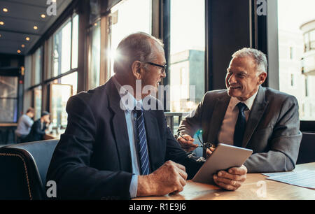 Deux hommes d'affaires seniors ayant une réunion informelle au café. homme d'affaires tenant la tablette numérique parlant avec son partenaire masculin tout en étant assis à co moderne Banque D'Images