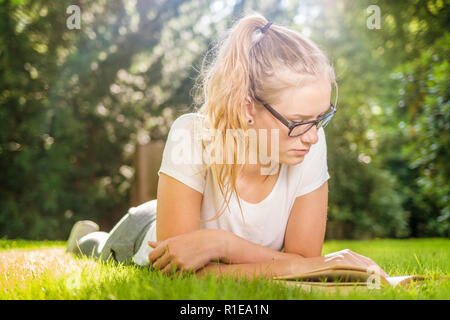 Une jeune femme avec des lunettes se trouve sur l'herbe dans le parc et l'air heureux dans un livre Banque D'Images