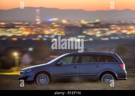 Déménagement voiture grise dans la nuit dans les prés verts sur fond de lumières de ville loin des immeubles et dark mountain ridge sous ciel clair au coucher du soleil. Transp Banque D'Images