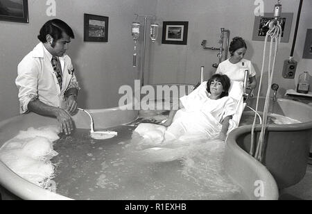 Années 1970, historiques, à l'intérieur d'une chambre dans un hôpital, la femelle adulte d'un patient allongé sur une civière métallique dans un grand bain à remous de l'eau moussante obtenir un traitement d'hydrothérapie, assisté d'une infirmière et d'un médecin traitant, Los Angeles, USA. Banque D'Images