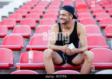 Smiling sporty man in headphones listening music assis sur les sièges du stade Banque D'Images