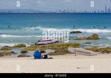 Lifeguard est assis sur la plage à l'ombre d'un parasol à regarder les surfeurs, avec des toits de la ville de Paradise Surf en arrière-plan. Banque D'Images