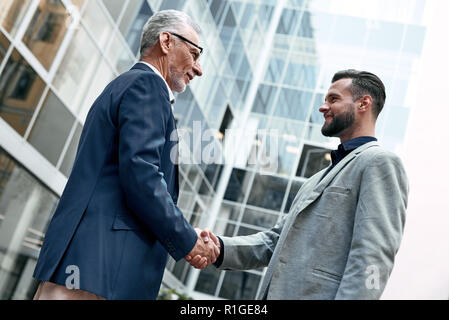 Réunion est démarré. Les jeunes et les cadres supérieurs smiling businessmen shaking hands ensemble tout en se tenant à l'extérieur sur l'arrière-plan de la ville Banque D'Images