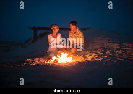 Happy young couple tenir la main et romantique du regard dans les yeux de chacun à côté d'un feu de camp sur la plage le soir. Banque D'Images