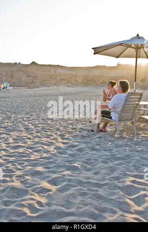 Mid-adult couple applaudir alors qu'il était assis sous un parasol sur une plage au soleil. Banque D'Images