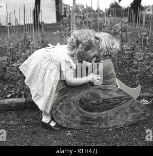 Années 1950, historiques, deux petites filles à l'extérieur par un jardin potager, un chuchotement à l'autre qui est assise sur une chaise berçante en bois canard. Banque D'Images