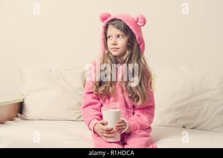 Portrait de jeune fille en pyjama rose doux et chaleureux. Fille 7 ans, blonde avec de longs cheveux bouclés dans le capot, avec une tasse de thé, à la voiture. La lumière d'arrière-plan h Banque D'Images