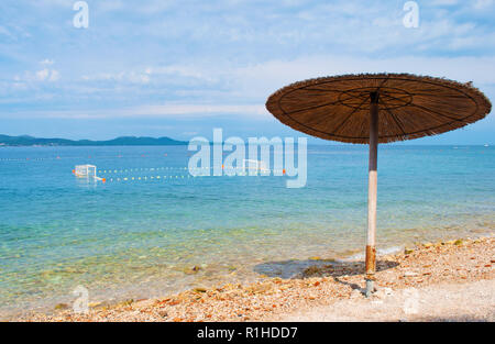 Un jaune parapluie de paille sur la plage de galets. La masse de water-polo avec un filet dans la mer bleu azur près de la côte. Le bleu ciel nuageux. Zadar, Croati Banque D'Images