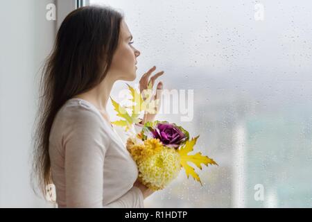 Bonjour l'automne. Jeune femme souriante avec automne bouquet de fleurs jaunes et orange, violet chou ornemental et feuilles d'érable. Femme près de la fenêtre Banque D'Images
