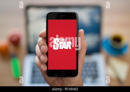 Un homme se penche sur son iPhone qui affiche le logo Daily Star, en restant assis devant son ordinateur 24 (usage éditorial uniquement). Banque D'Images