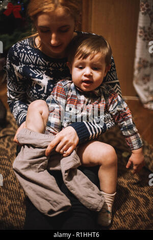 Mère s'habiller son enfant assis sur le plancher à la maison durant les fêtes de Noël Banque D'Images