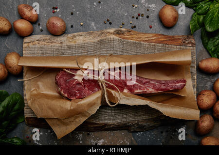 Bifteck de faux-filet cru enveloppés dans du papier sulfurisé sur planche de bois avec des épices et pommes de terre Banque D'Images