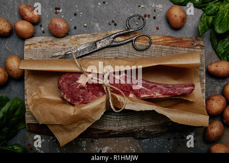 Vue de dessus de bifteck de faux-filet cru enveloppés dans du papier sulfurisé sur planche de bois avec des épices et pommes de terre Banque D'Images