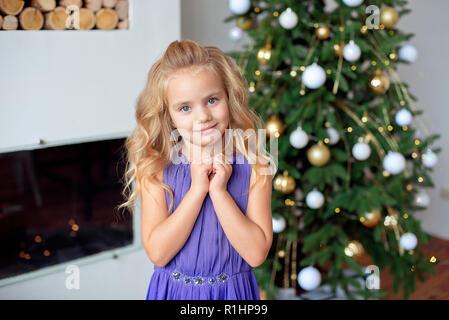 Happy little girl en violet robe de luxe détient près de mains visage.avec lumières colorées et l'arbre de Noël sur l'arrière-plan. Jours fériés, Noël, nouvel an, x Banque D'Images