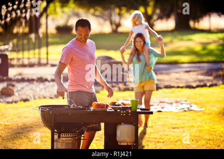 Jeune fille assise sur les épaules de sa mère pendant que son père cuisiniers sur un barbecue. Banque D'Images