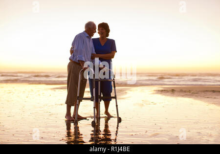 Man et une infirmière avec une aide à la marche sur une plage au coucher du soleil. Banque D'Images