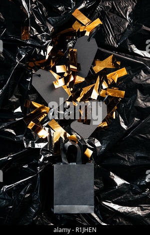 Vue de dessus du panier de papier vierge avec des étiquettes de prix et les confettis d'or noir sur fond noir, papier d'emballage concept vendredi Banque D'Images