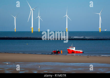 Les pêcheurs à l'aide d'un tracteur et remorque rouge pour récupérer un bateau de pêche sur la plage de Redcar North Yorkshire UK avec la ferme éolienne derrière Banque D'Images