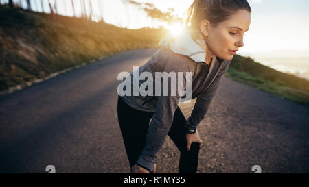 Coureuse en faisant une pause pendant son jogging du matin debout sur une rue avec le soleil en arrière-plan. Athlète femme se détendre après son repos d'entraînement Banque D'Images