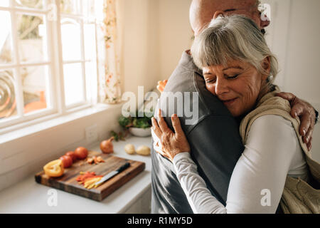 Vue latérale d'un couple de personnes âgées serrant les uns les autres à la maison. Senior woman embracing son mari aux yeux clos debout dans la cuisine. Banque D'Images