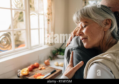 Senior couple at home enlacés debout dans la cuisine. Senior woman donnant une accolade chaleureuse à son mari dans l'expression de son amour. Banque D'Images