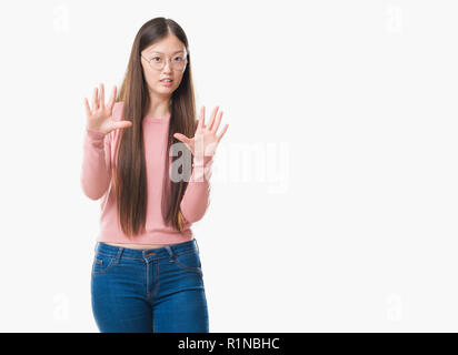 Jeune femme chinoise sur fond isolé portant des lunettes peur et terrifié par la peur d'arrêt expression geste avec les mains, criant sous le choc. Pan Banque D'Images