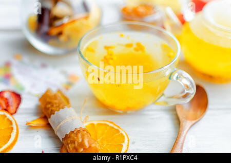 L'argousier thé dans la théière et tasse en verre et de nombreux fruits divers et de pastilles, plaquettes sur le petit déjeuner sur la table en bois blanc, lumière naturelle, l'accent selektive Banque D'Images