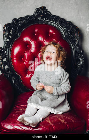 Rire fille aux cheveux bouclés en robe rouge gris assis en fauteuil. Banque D'Images