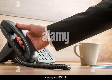 Main d'un businessman holding black téléphone fixe combiné pour le récupérer à partir de la base pour répondre à un appel téléphonique. Tasse à café avec à côté de lui sur woode Banque D'Images