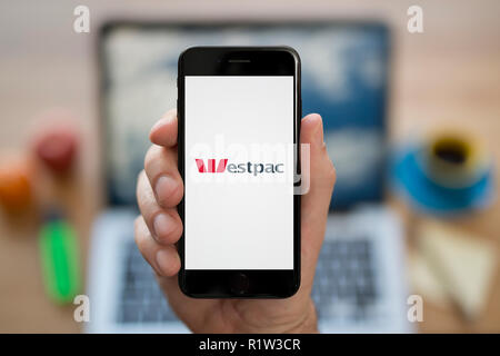 Un homme se penche sur son iPhone qui affiche le logo de Westpac, tandis qu'assis à son bureau de l'ordinateur (usage éditorial uniquement). Banque D'Images