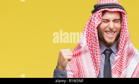 Beau jeune homme arabe aux cheveux longs portant keffieh sur fond isolé très heureux et excité de faire geste gagnant avec bras levés, SMIL Banque D'Images