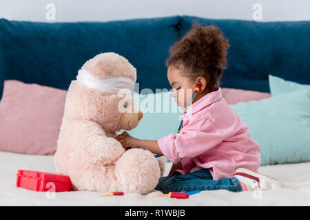 Peu d'enfants afro-américains dans dossier rose jouant le médecin avec ours Banque D'Images