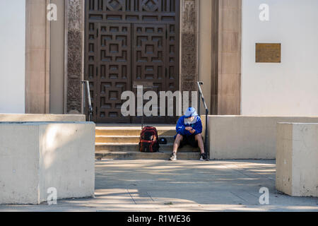 Sans-abri d'un seul homme est assis sur les marches de la bibliothèque de la ville avec son sac à dos et vêtu d'un poncho et des courts-circuits. Banque D'Images