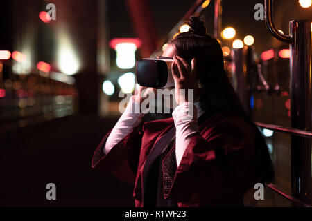 Fille asiatique attrayant à l'aide de casque de réalité virtuelle sur la rue en soirée, ville d'avenir concept Banque D'Images